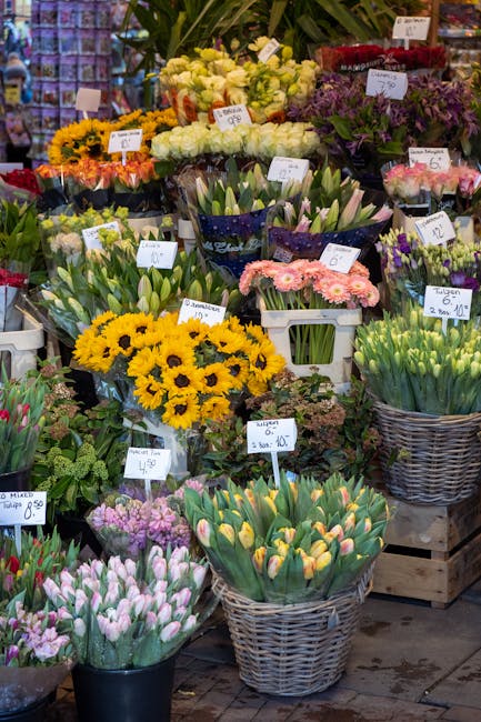 A vibrant flower display at a local market featuring a variety of fresh flowers including yellow sunflowers, white roses, pink tulips, purple irises, and tulips in shades of pink and yellow arranged i