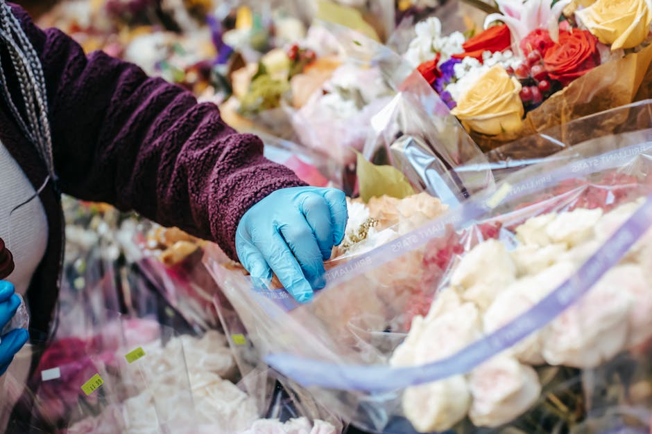 A person wearing a dark purple textured sweater and blue gloves selecting a bouquet of fresh flowers from a display at a florist, with multiple floral arrangements wrapped in clear plastic and paper i