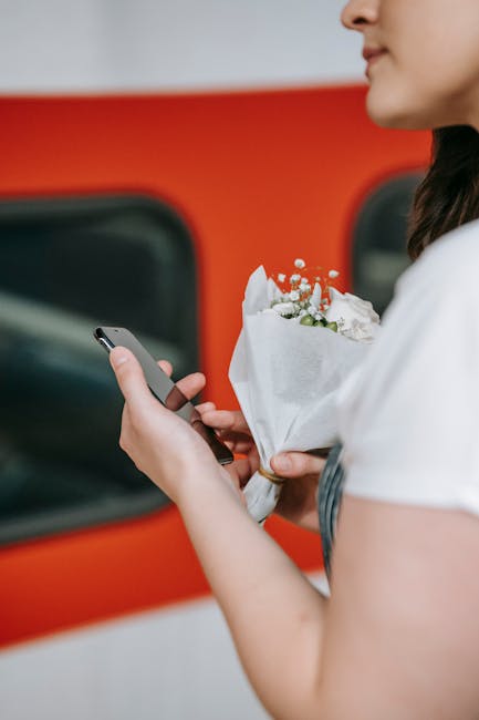 Close-up of a woman holding a small bouquet of fresh white and green flowers wrapped in white tissue paper, standing beside a red vehicle with a dark window. She is using a smartphone with her other h