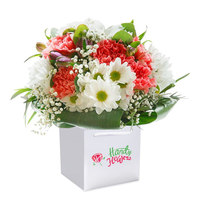 Presented upright in a sparkling white, square gift bag, this bouquet offers a vibrant tableau sure to catch the eye of anyone in New Southgate. Coral and peach carnations, with their signature ruffled petals and inviting colour, are framed by snowy-white daisy chrysanthemums topped with lively green centers, together forming a cheerful medley. Throughout the bouquet, delicate stems of white gypsophila add softness and depth, gently diffusing light for a cloud-like effect, while glossy dark-green foliage wraps around the base, providing stability and visual richness reminiscent of local parks near Brunswick Avenue. The addition of closed, purple-tinged buds offers subtle depth and a sense of graceful anticipation, echoing New Southgate's spirit of quiet growth. The white gift bag, designed for easy handling and chic presentation, sports refined ribbon handles-ideal for hand-delivery to homes or offices within the community. The arrangement's balanced, full silhouette and striking mix of hues are heightened by gentle, professional lighting, allowing every bloom's vibrancy to shine. Each blossom is flawlessly fresh, echoing the commitment to quality for which local florists in New Southgate are known. Suitable for occasions such as birthdays, anniversaries, or as a bright 'thinking of you' gift, this floral creation captures the essence of New Southgate's warmth and neighbourly connection.