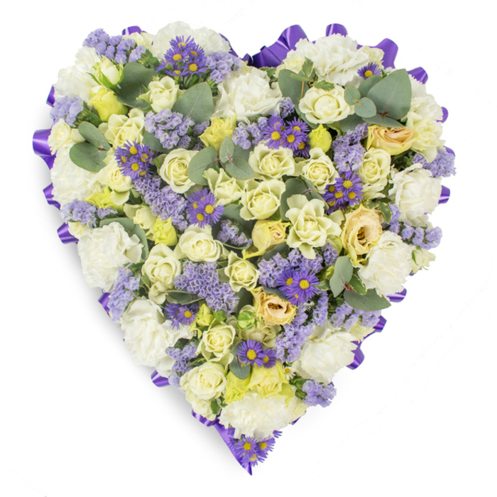 This photograph captures a lovingly assembled heart-shaped funeral tribute, set on a pristine white background so the floral details read with clear, gentle intent. The outline is neatly finished with a continuous band of rich purple satin ribbon, looped and folded to form an elegant border that contains the dense bed of blooms within. The primary tonality is soft: clusters of cream and pale yellow roses - their petals smooth and closely layered - create a tactile centre, their subtle gradations of colour suggesting warmth and comfort. Scattered among the roses are abundant white carnations, their ruffled texture building volume and a classic sympathy feel. Interspersed throughout are wisps of lavender statice, lending a delicate, papery texture and a subdued lilac tone, while small, vivid purple aster-like flowers with bright yellow centres act as focused accents that draw the eye across the heart. The composition is breathed upon by rounded, silvery-blue eucalyptus leaves, which both punctuate the arrangement visually and imply a clean, herbal scent. The soft, even lighting used in the studio photograph emphasizes the layered depth of the flowers without dramatic shadows, presenting the tribute as serene and composed. This type of arrangement is frequently chosen for wakes, funeral services or grave-side remembrances around New Southgate, Arnos Grove and Bowes Park, where families seek a compassionate, enduring expression of love and memory, rendered with considered craftsmanship and quiet dignity.