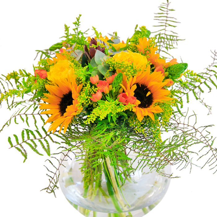 Bathed in gentle sunlight, this luxurious arrangement echoes the late summer charm of New Southgate's leafy avenues. The vivid tableau begins with large sunflowers-petals like rays of sunshine radiate from rich espresso centers, bringing a sense of outdoorsy energy to the composition. Nestled closely are rosettes of yellow garden roses, their layered petals tightly wound in spirals hinting at careful craftsmanship and a love for botanical detail. Highlights of bright red Hypericum berries glisten amid waves of fresh, green foliage; their clustered forms bring a playful vibrancy reminiscent of market bouquets found near Osidge Lane. Cascading ferns and whisps of goldenrod add airy movement-each frond and blossom playing with the light, dancing gently like summer breezes near New Southgate Library's gardens. Eucalyptus leaves add a cool, bluish touch, their distinctive form peeking throughout, creating visual depth and rustic charm. Set in a pristine, round glass vase, the stems weave together beneath the surface, their bright green colors magnified by clear water. The bouquet exudes a delicate floral scent, subtle yet lively, blending the sweetness of roses and the earthiness of sunflowers. Perfect for commemorating milestones or spreading cheer to homes and offices in New Southgate, this arrangement is an enchanting celebration of happiness, crafted with the personalized flair only a dedicated local florist can provide.