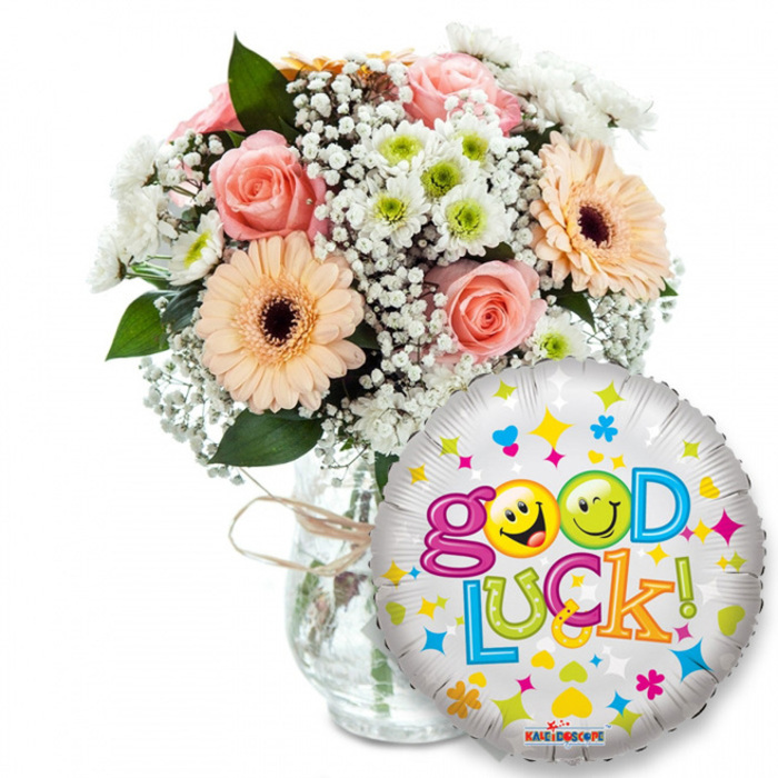 A bright, supportive arrangement captured on a clean white field: a full floral bouquet in a clear glass vase stands slightly left, finished with a rustic twine bow that suggests an artisan touch. The bouquet blends warm peachy-orange gerbera daisies with dark central discs, gentle pale-pink roses whose petals fold in soft spirals, generous sprays of tiny white baby's breath (Gypsophila) and compact clusters of white and lime-green chrysanthemums that add freshness and texture. Deep green foliage threads between blooms, anchoring the pastel palette and lending natural depth. Prominently placed to the right and overlapping the lower right of the vase, a round silver metallic foil balloon sparkles with a scatter of colourful stars, hearts and abstract shapes in blue, pink, yellow and green. The headline Good Luck! is rendered in lively, multi-coloured letters; the two 'o's in Good are playful yellow smiley faces - one giving a wink, the other sticking its tongue out - which makes the message feel intimate and upbeat. You can almost sense the contrast in textures: the velvety softness of roses and gerberas against the crisp, papery chrysanthemums and the slick, reflective sheen of the balloon, topped by the subtle KALEIDOSCOPE logo at the balloon's base. Bathed in soft, neutral light that highlights the pastel tones, this composition radiates encouragement - ideal for sending best wishes for exams, a new job, graduations or a warm farewell. As a local florist familiar with New Southgate and nearby Brunswick Park, I picture delivering this to a front door on Friern Barnet Road, an immediate, heartfelt lift for any milestone.