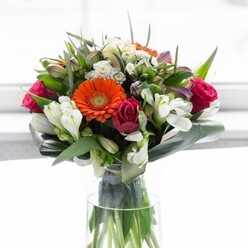This exquisite floral arrangement, crafted by seasoned florists in New Southgate, is a lesson in balance, artistry, and emotion. Clusters of creamy peach and dynamic orange gerbera daisies form a gentle, glowing crown that sparkles in diffused natural light, evoking sunrise over the leafy lanes of neighboring Oakleigh Park. Rich fuchsia roses anchor the bouquet, their velvety petals swirling tightly-suggesting both passion and sophistication. Pristine white alstroemeria and delicate spray roses interlace throughout, shimmering with a subtle luminescence that adds brightness and layers to the ensemble. The surrounding greenery-bold, glossy aspidistra leaves gracefully curved around the base and fine, feathery greens filling in-grounds the vivid florals in a sense of natural abundance. Every stem is presented within a clear, high-sided glass vase that highlights the living, green architecture beneath, while the play of soft sunlight keeps the focus on the vibrant blooms above. Subtle aromas mingle: a trace of gentle lily, the sweetness of rose, the clean essence of freshly cut greenery. Together, these create a joyful, optimistic mood that suits celebrations, heartfelt thank-yous, or fresh beginnings in a local home. Each detail hints at the expertise of Flower Delivery New Southgate-where creativity blends with trusted local service, and every bouquet carries a sense of personal connection between florist and community.